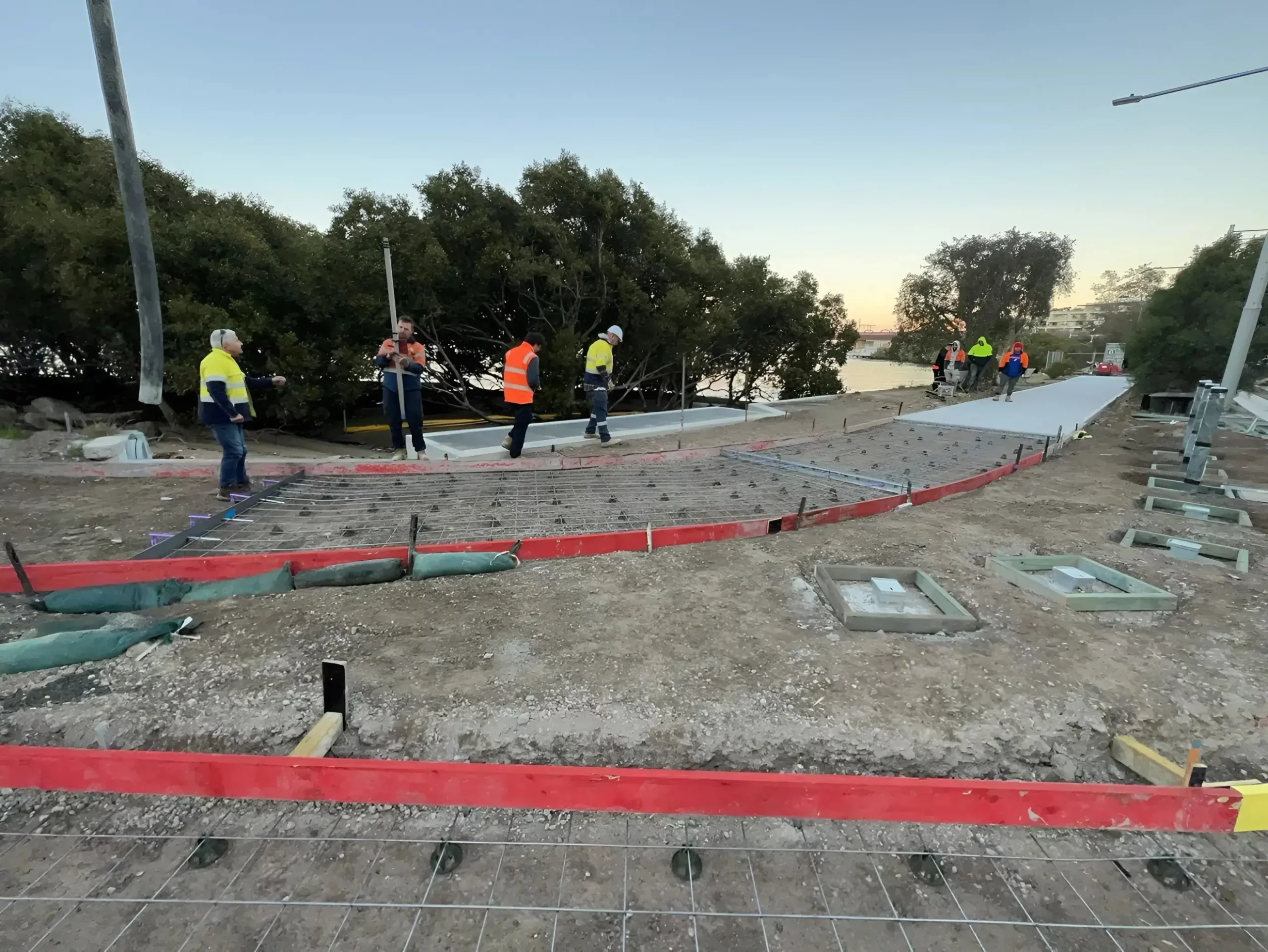 Several construction workers wearing high-visibility clothing and hard hats are working on a site with concrete paths and metal reinforcements. The site is in a semi-rural area near trees and water, with the sun setting in the background.