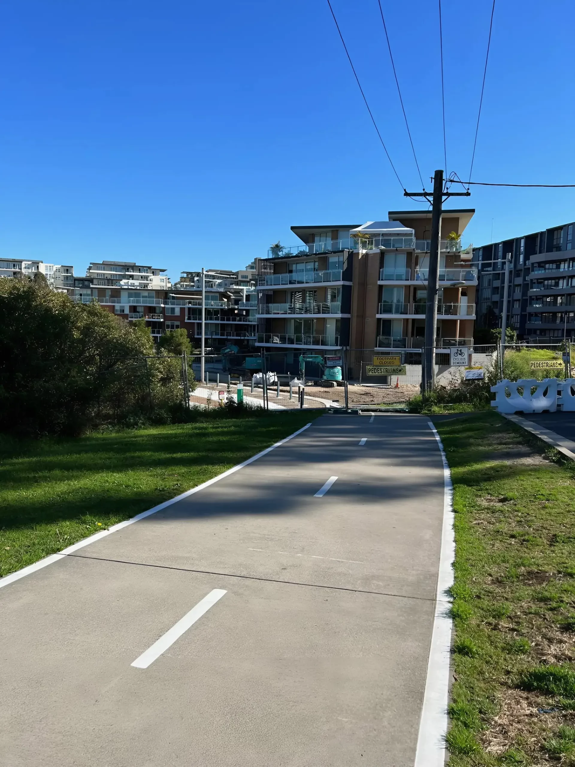 A paved pathway with white lane markings under a clear blue sky cuts through a green grassy area. It leads towards modern apartment buildings in the background. Power lines and a construction site are visible on the right side.