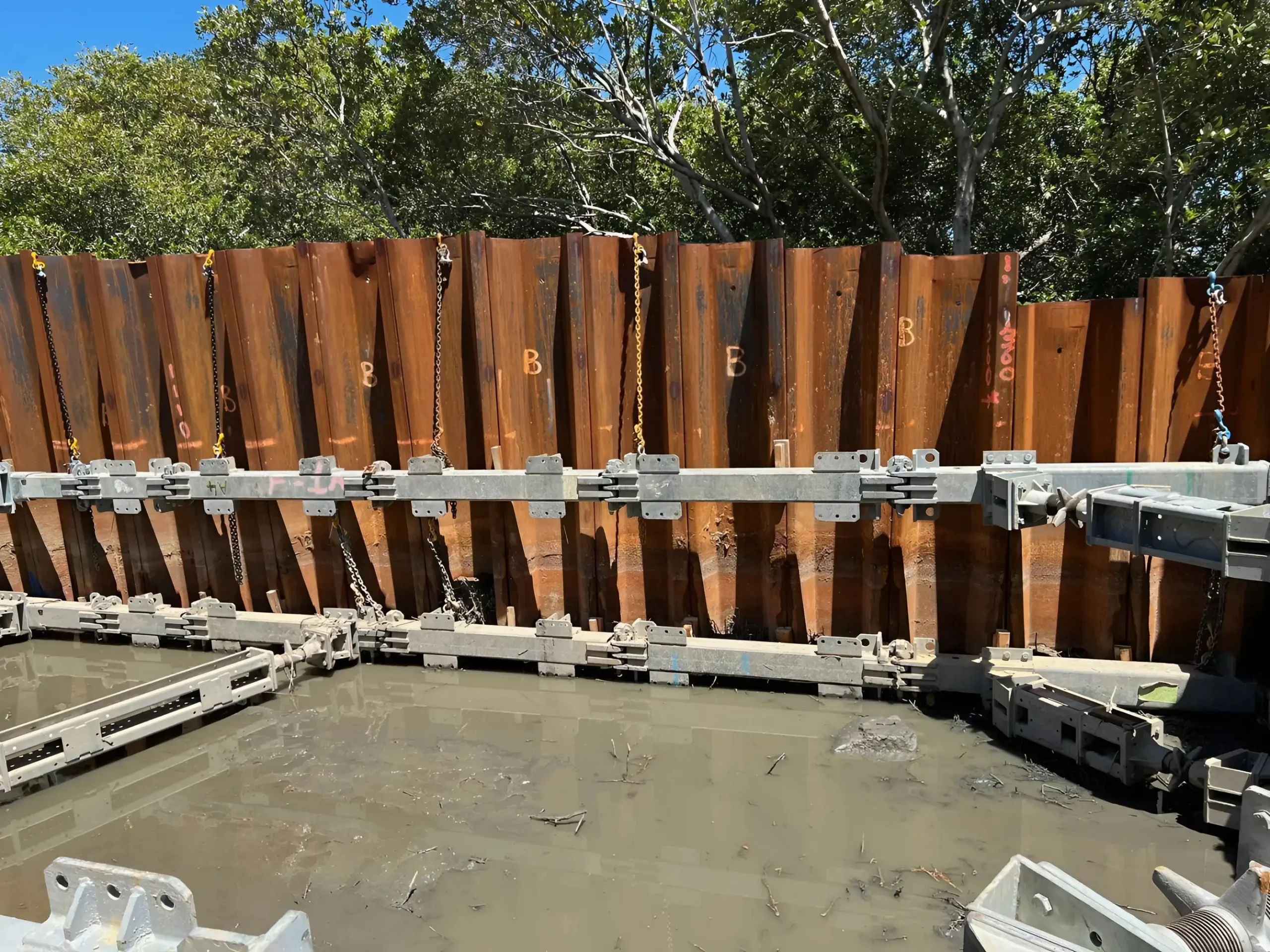 A construction site with large rusty steel sheet piles driven into the muddy ground, forming a retaining wall. Metal supports and braces are attached horizontally across the piles. Trees and greenery are visible in the background under a clear blue sky.