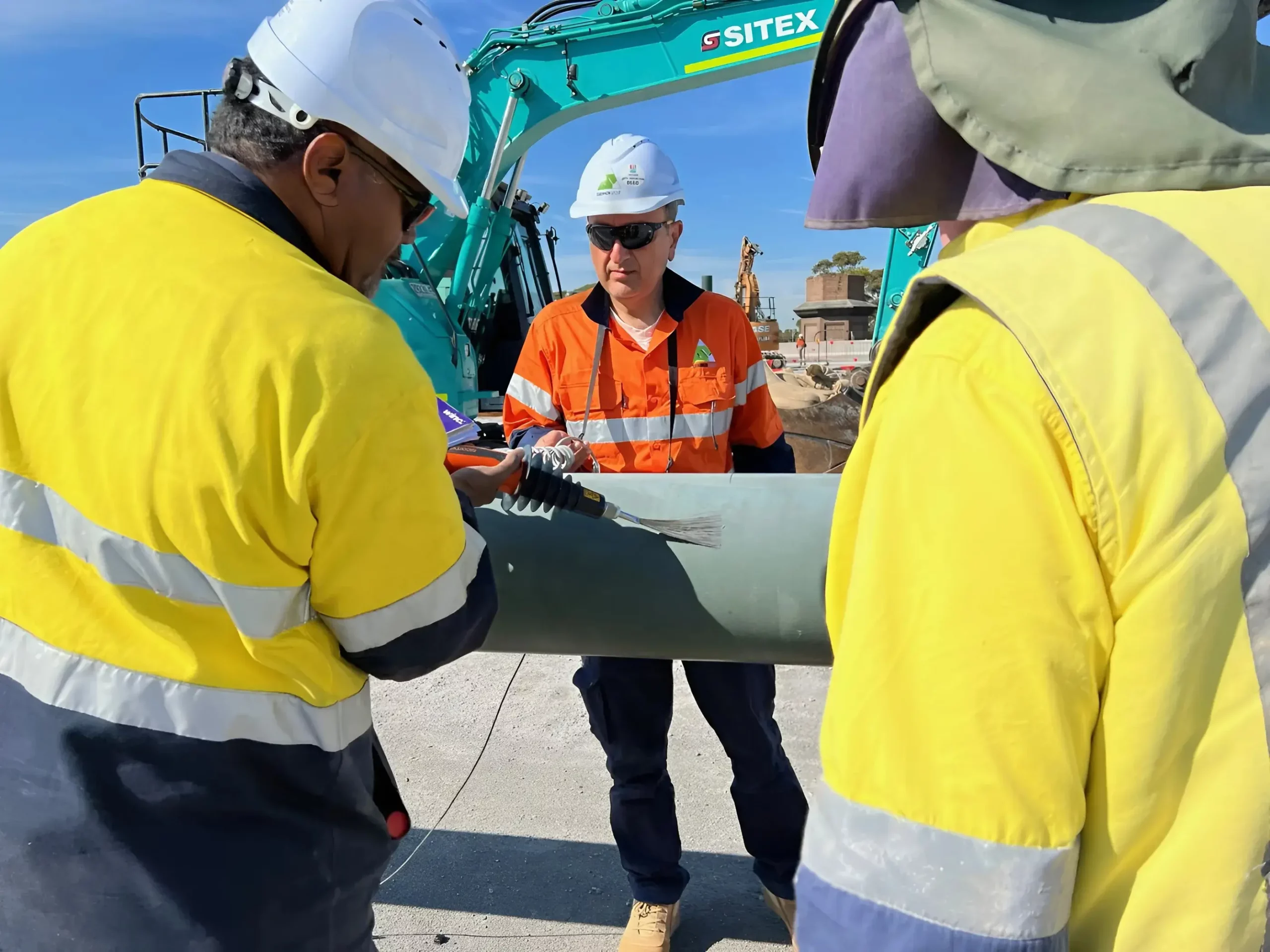 Three construction workers, wearing safety gear and helmets, work together on a project at a construction site. One worker in an orange vest operates a tool on a large metal pipe, while the other two in yellow vests assist. Excavator in the background.