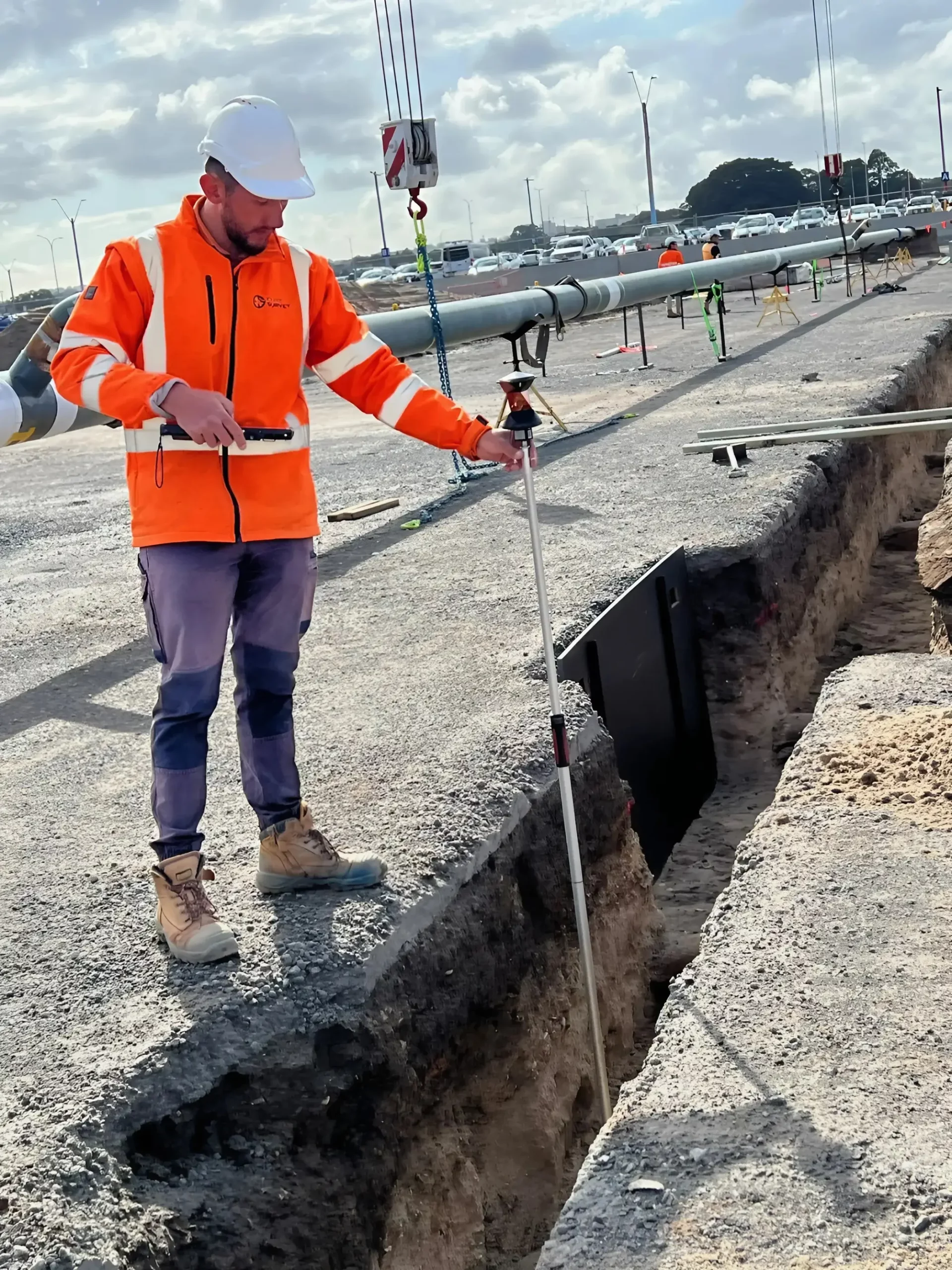 A construction worker wearing an orange high-visibility jacket, hard hat, and boots stands beside a trench at a construction site. The worker holds a measuring rod and uses it to measure the depth of the trench, which is lined with safety barriers.