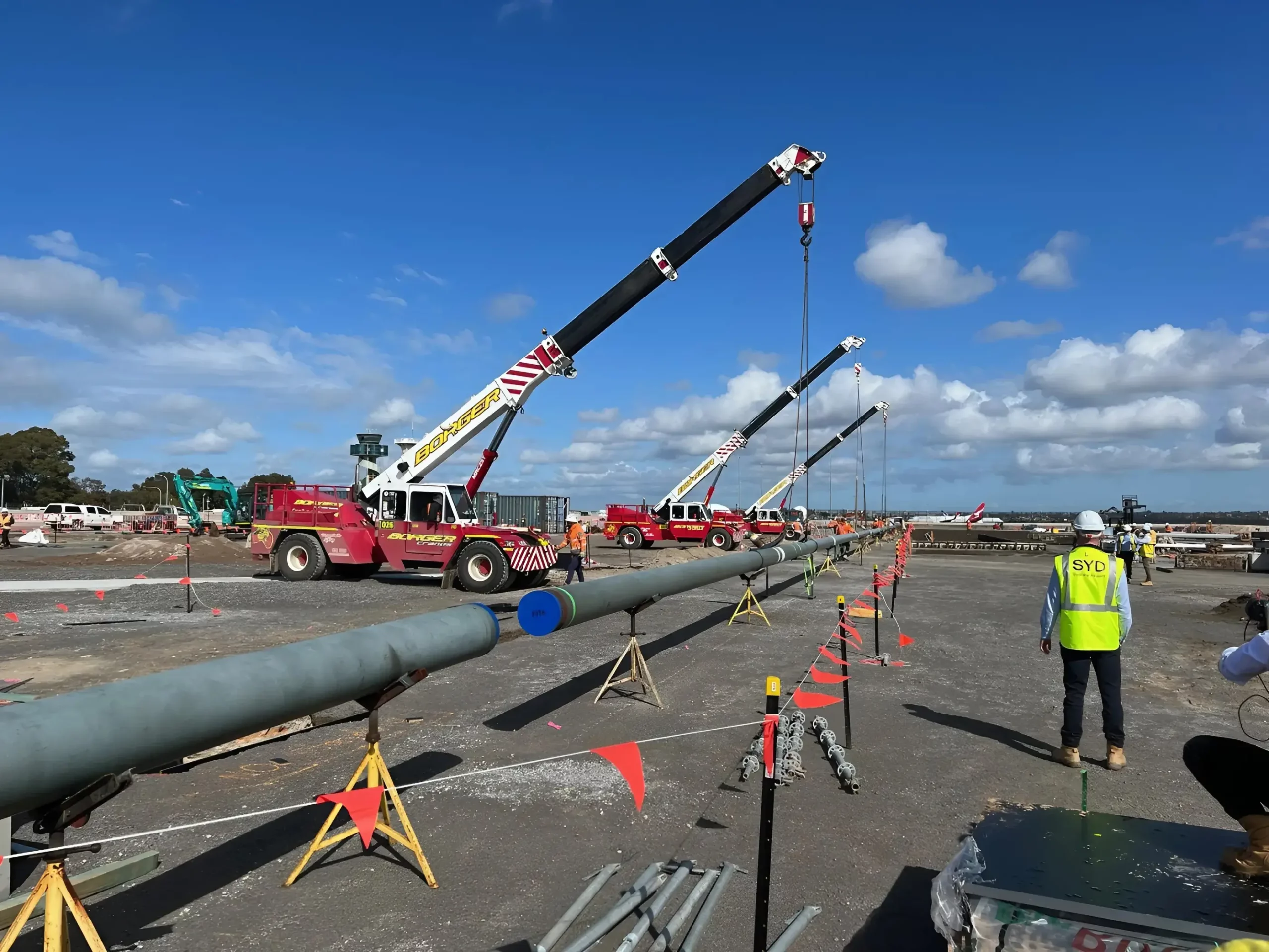 Construction site with multiple cranes lifting large pipes. Workers, including one in a high-visibility vest labeled “SYC,” are overseeing the operation. Traffic cones and barricades outline the work area. The sky is clear with a few clouds.