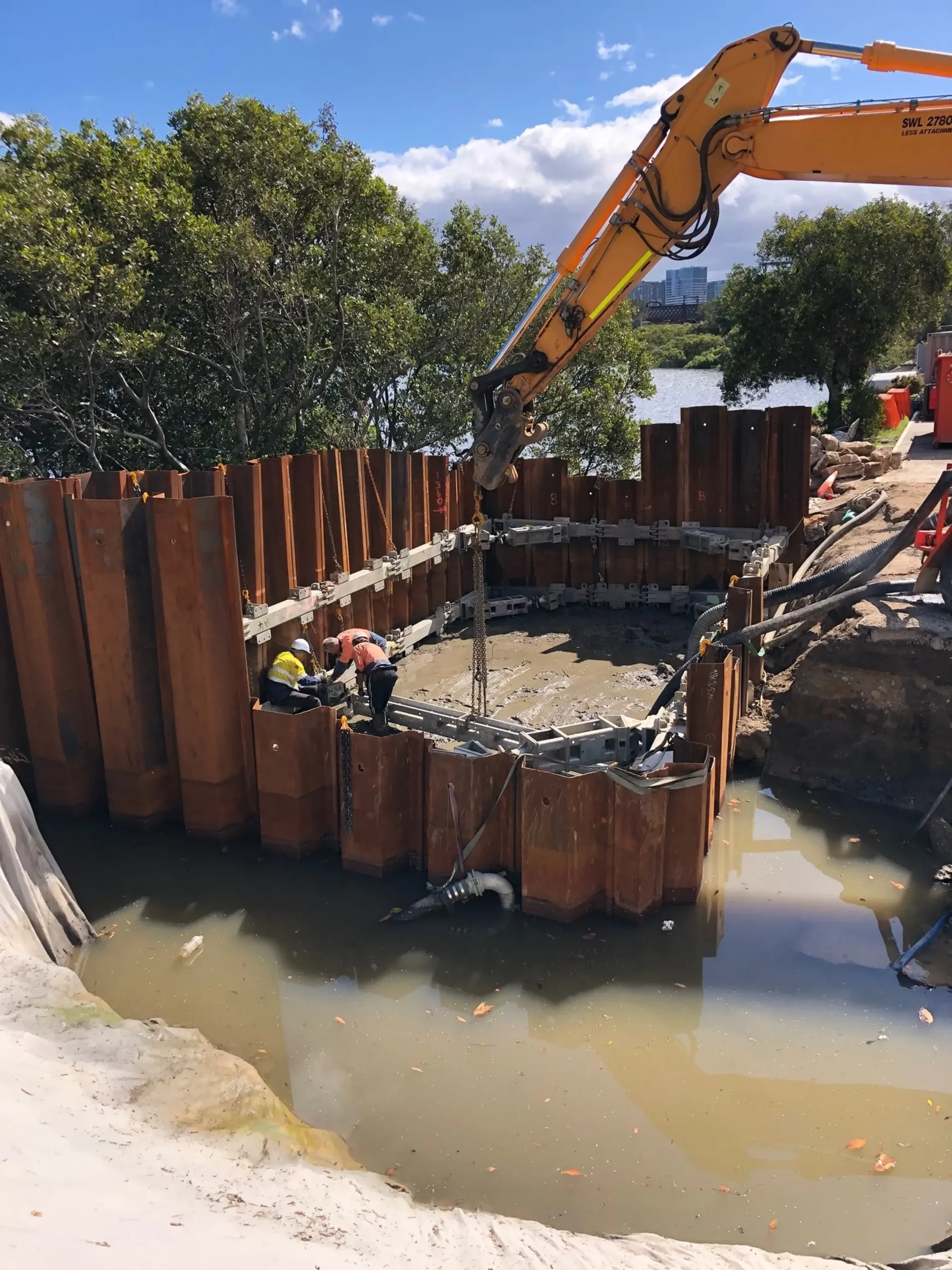 Construction workers inside a large, steel-reinforced excavation site surrounded by wooden beams. An excavator with a long arm assists in the work. The setting is near a body of water with trees in the background, suggesting a waterfront construction project.