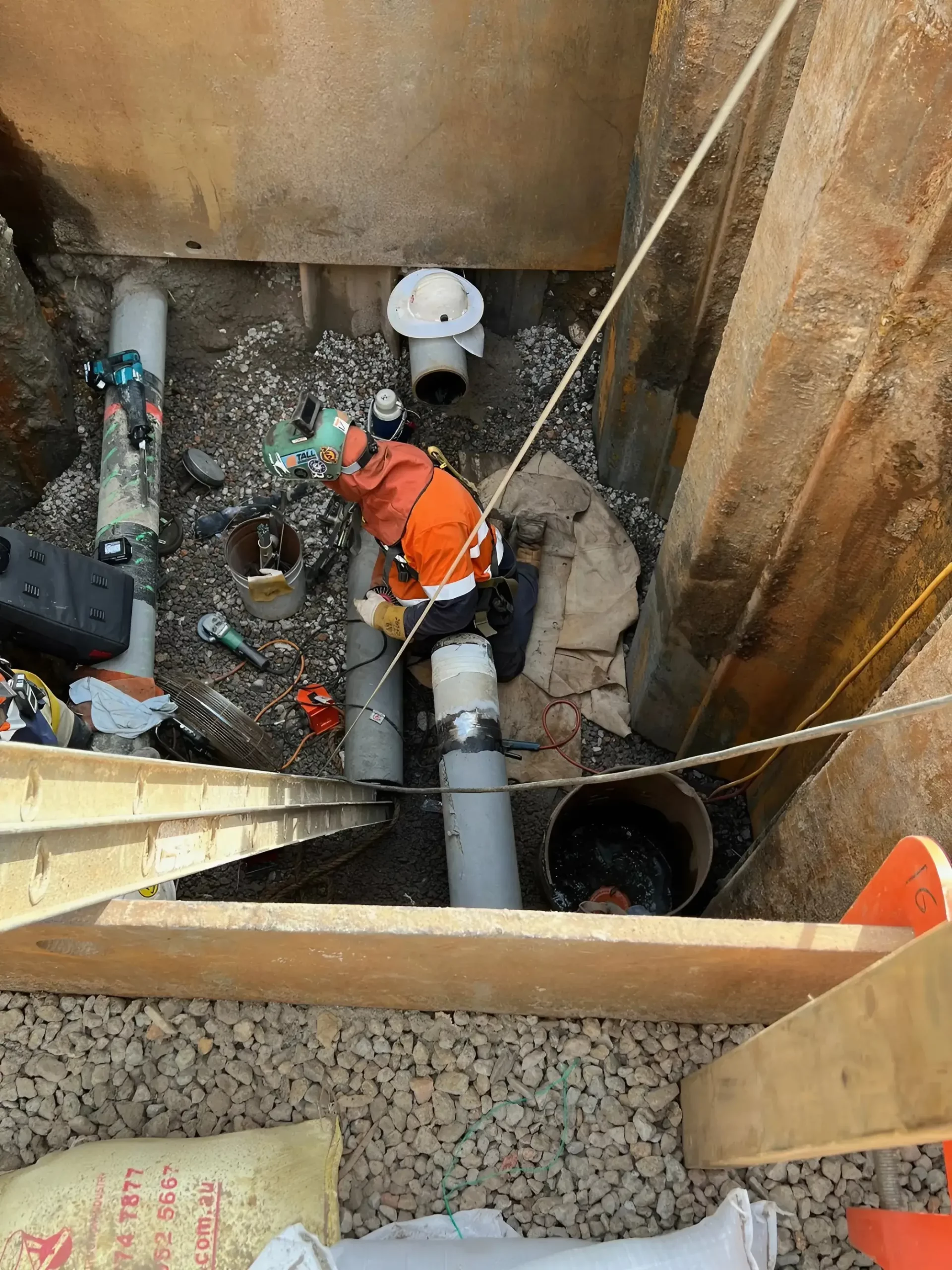 A worker in an orange jacket and hard hat is seen from above in a construction trench. They are surrounded by large pipes, tools, and construction materials, working on a pipe connection. The trench walls are supported by wooden and metal shoring.