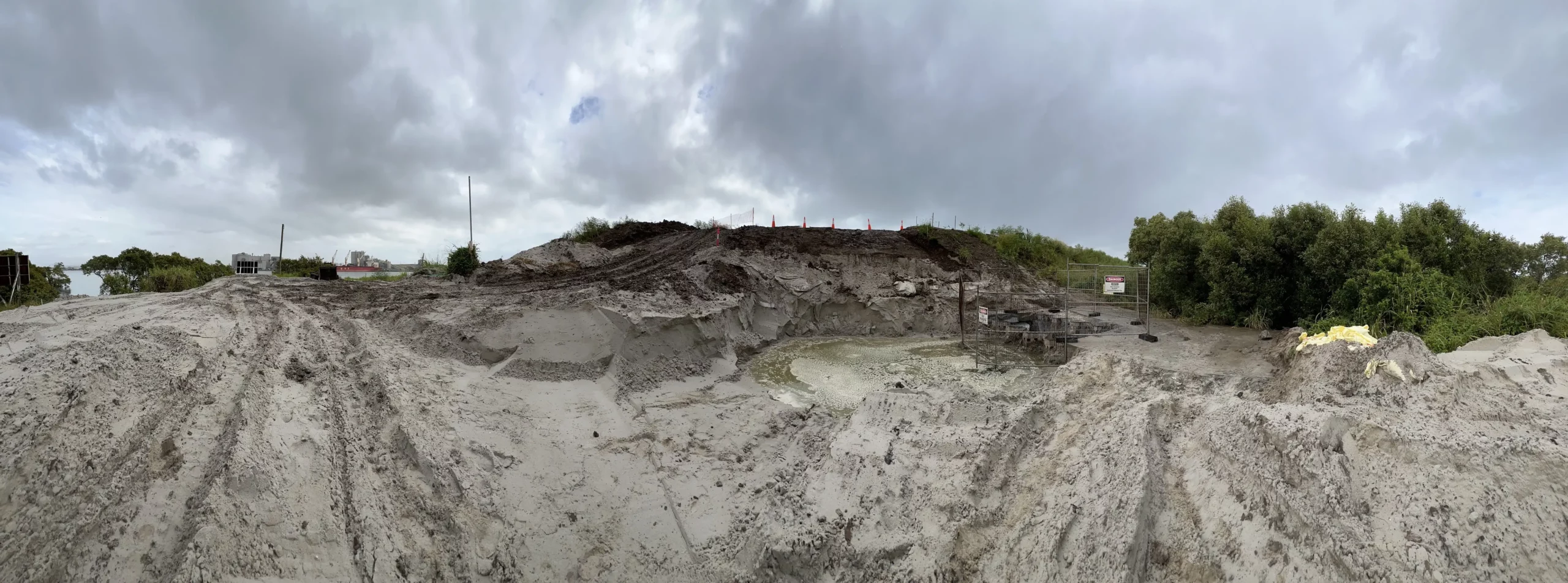 Panoramic view of a muddy construction site with large dirt piles and machinery. The sky is overcast with gray clouds, and sparse vegetation is visible in the background.