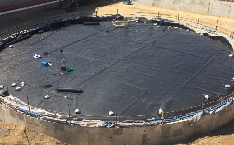 A large, circular concrete structure is under construction, lined with black plastic material. Various tools and equipment are scattered on the surface, and the area is enclosed with safety barriers. The surrounding ground is sandy and appears at an early construction stage.